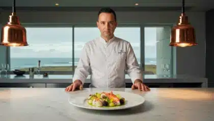 Le chef Mathieu Guibert posant derrière une assiette gastronomique dans son restaurant avec vue sur l’océan, sous deux lampes en cuivre.