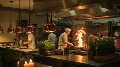 Cuisine professionnelle avec des chefs en plein service, l’un d’eux faisant flamber une poêle sous la hotte.