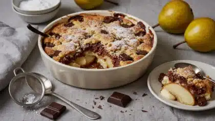 Crumble poire-chocolat doré et fondant de Cyril Lignac, saupoudré de sucre glace, servi avec des morceaux de chocolat noir et des poires fraîches.