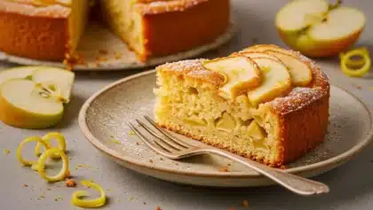 Part de gâteau moelleux madeleine-pommes servie dans une assiette, décorée de zestes de citron et de tranches de pomme dorées.