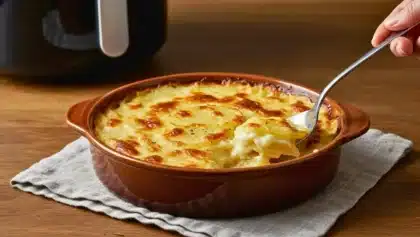 Gratin dauphinois doré dans un plat en céramique, avec une cuillère prélevant une portion fondante, posé sur un torchon devant un Airfryer sur une table en bois.