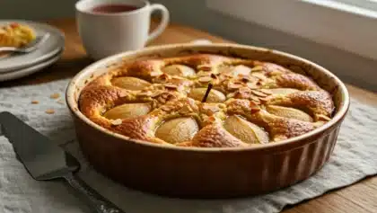Gâteau minute poire–amande dans un moule rond en céramique, doré et garni d’amandes effilées, avec des poires entières visibles à la surface, posé sur un linge clair avec une tasse de thé à l’arrière-plan.