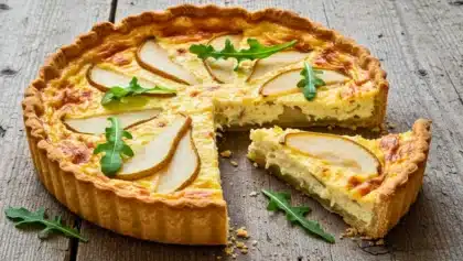Quiche dorée garnie de tranches fines de poire et de feuilles de roquette, avec une part découpée sur une planche en bois.