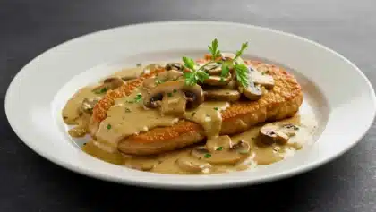 Escalope de veau nappée de sauce à la crème et aux champignons, garnie de persil frais, servie dans une assiette blanche sur fond sombre.