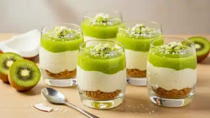 Verrines en trois couches avec biscuit émietté, mousse coco blanche et coulis de kiwi vert, décorées de dés de kiwi et de coco râpée, posées sur une table en bois.