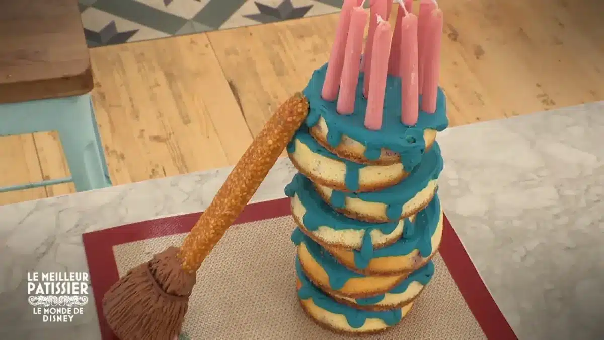 Gâteau à étages recouvert d’un glaçage bleu, décoré de bougies roses et d’un manche en biscuit évoquant un balai magique, inspiré du monde de Disney dans Le Meilleur Pâtissier.
