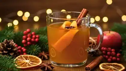 Verre de jus de pomme chaud aux épices avec bâton de cannelle, écorce d’orange et étoile de badiane, décoré d’éléments de Noël sur une table en bois.