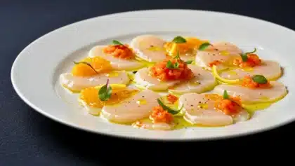 Assiette de carpaccio de Saint-Jacques disposées en rosace, garnies de suprêmes d’orange, de condiment orangé, de micro-pousses et d’un filet d’huile d’olive sur fond sombre.
