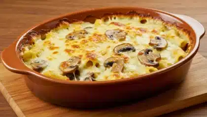Gratin de pâtes crémeux aux champignons dans un plat en céramique, avec fromage fondu doré, tranches de champignons visibles et surface gratinée, posé sur une planche en bois.
