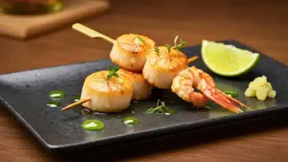 Brochette de Saint-Jacques et crevettes poêlées, nappées de beurre gingembre-soja, servie avec citron vert et herbes fraîches sur assiette noire.
