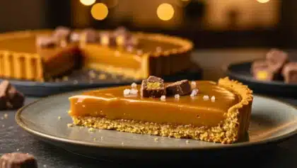 Part de tarte au Daim maison avec base biscuitée, caramel fondant et ganache au chocolat au lait, décorée de morceaux de Daim et de fleur de sel, servie sur une assiette.