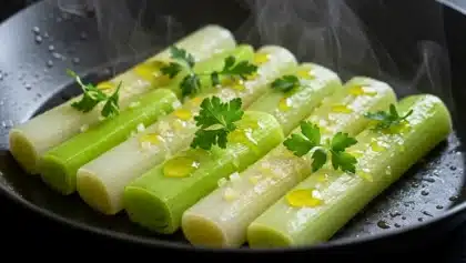 Tronçons de poireaux cuits doucement dans une poêle, nappés d’huile, parsemés de fleur de sel et de persil frais, avec vapeur visible.