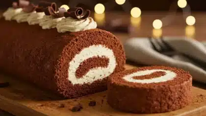 Gâteau roulé au chocolat garni de chantilly, tranché, décoré de copeaux de chocolat et de crème, présenté sur une planche en bois avec un fond de lumières floues.