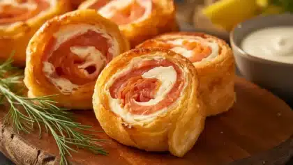 Roulés feuilletés apéritifs au saumon fumé et fromage frais, coupés en spirales dorées, disposés sur une planche en bois avec herbes fraîches.
