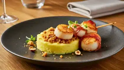 Brochette de Saint-Jacques grillées au lard servie sur purée de Ratte du Touquet avec crumble de noisettes dans une assiette noire.