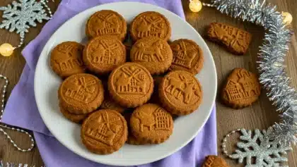 Assiette de spéculoos maison dorés, aux motifs traditionnels, présentée sur une table décorée d’éléments de Noël comme des guirlandes et flocons argentés.
