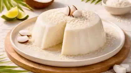 Flan coco blanc et fondant, coupé pour révéler son intérieur crémeux, présenté sur une assiette en céramique posée sur une table en bois clair avec décor tropical discret.