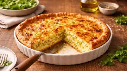 Une quiche aux poireaux dorée et fondante, présentée en vue à 45 degrés, avec une part nette découpée dans un plat blanc sur une table rustique soigneusement dressée.