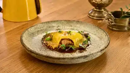 Une raviole dorée farcie au foie gras, dressée au centre d’une assiette rustique avec sauce brillante et jeunes pousses, sur une table en bois.